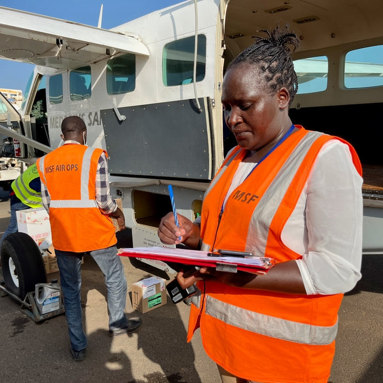 Person in an orange safety vest with a clipboard near a small aircraft at an airport.
