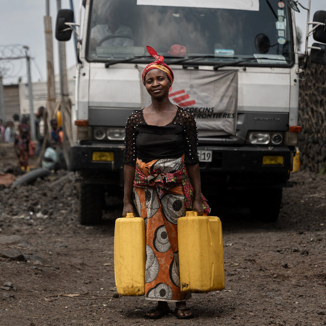 Woman carrying yellow jerry cans in an urban setting with a bus in the background