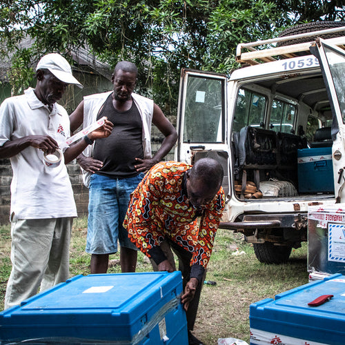 Three men standing near a van with open doors, surrounded by blue boxes on a grassy area.